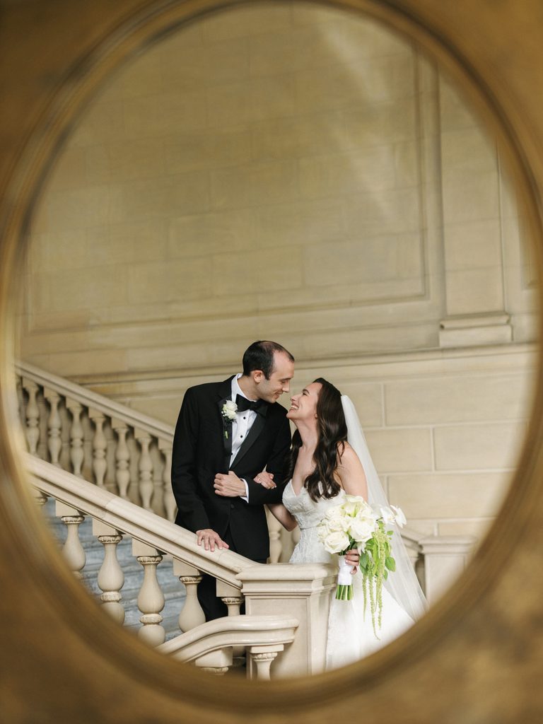 Luxury wedding couple sharing a romantic candid moment on a stone stairway captured through a beautiful gold framed mirror