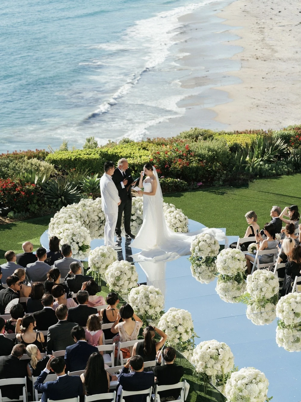 Outdoor wedding ceremony overlooking the ocean, with seated guests and white flowers.