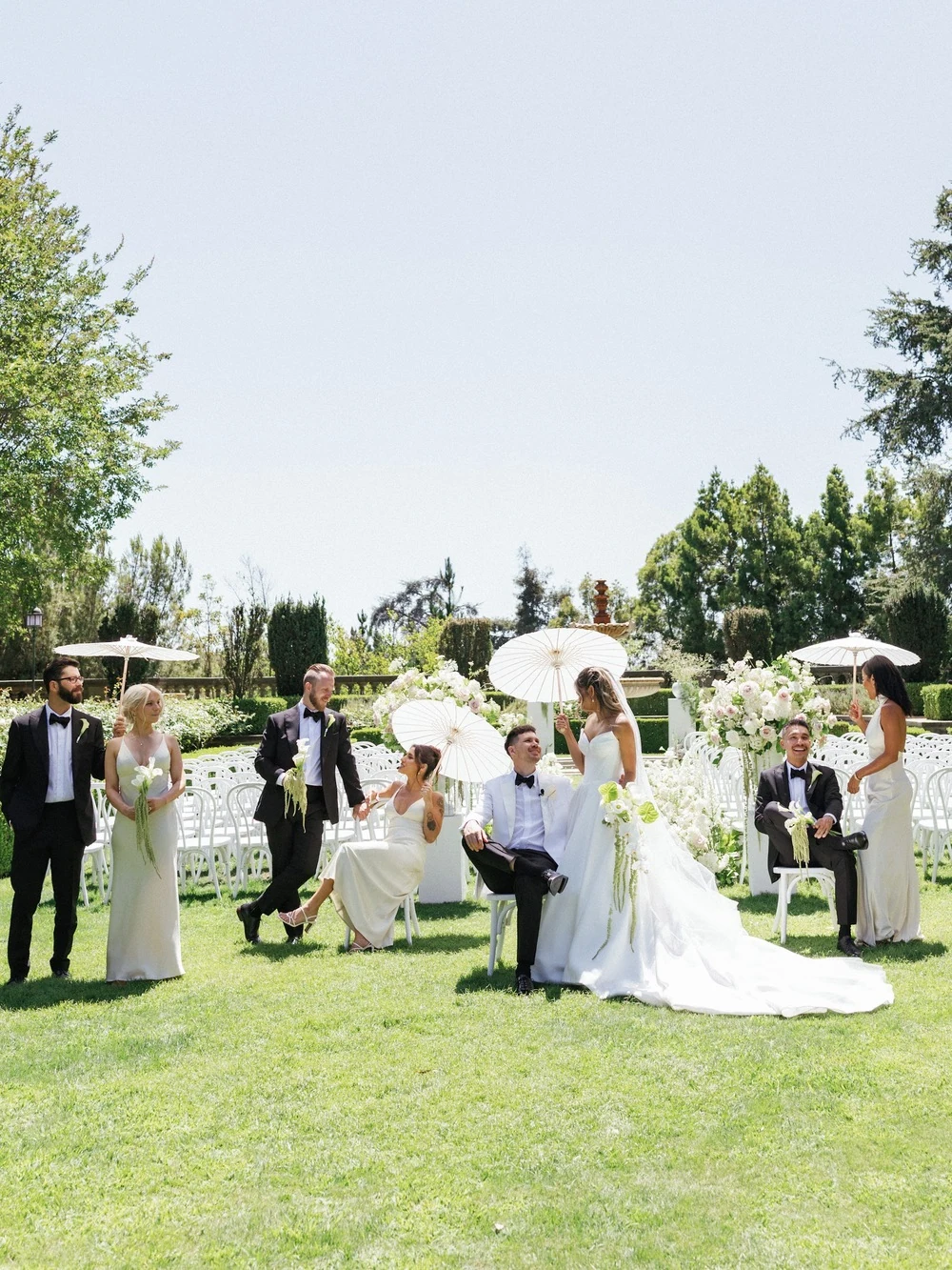 Wedding party gathered on a lawn with white parasols.